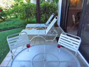 Picture of the lanai with the table for four in the forground only showing two seats and two glasses of wine. The two lounge chairs and tropical foliage are in the background.